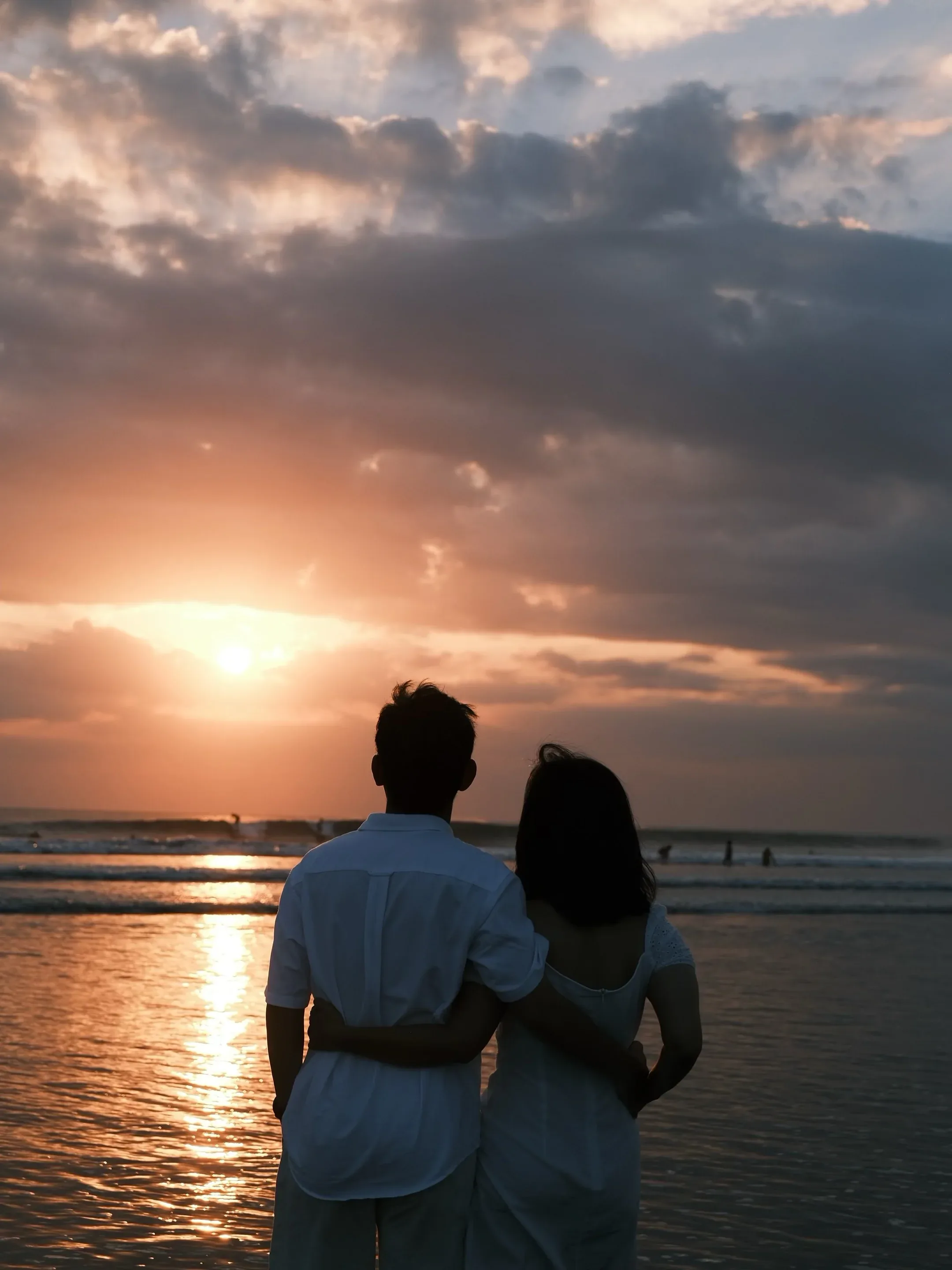 The couple from behind, his arm around her, watching the sun drop into the water under a wide cloudy sky.