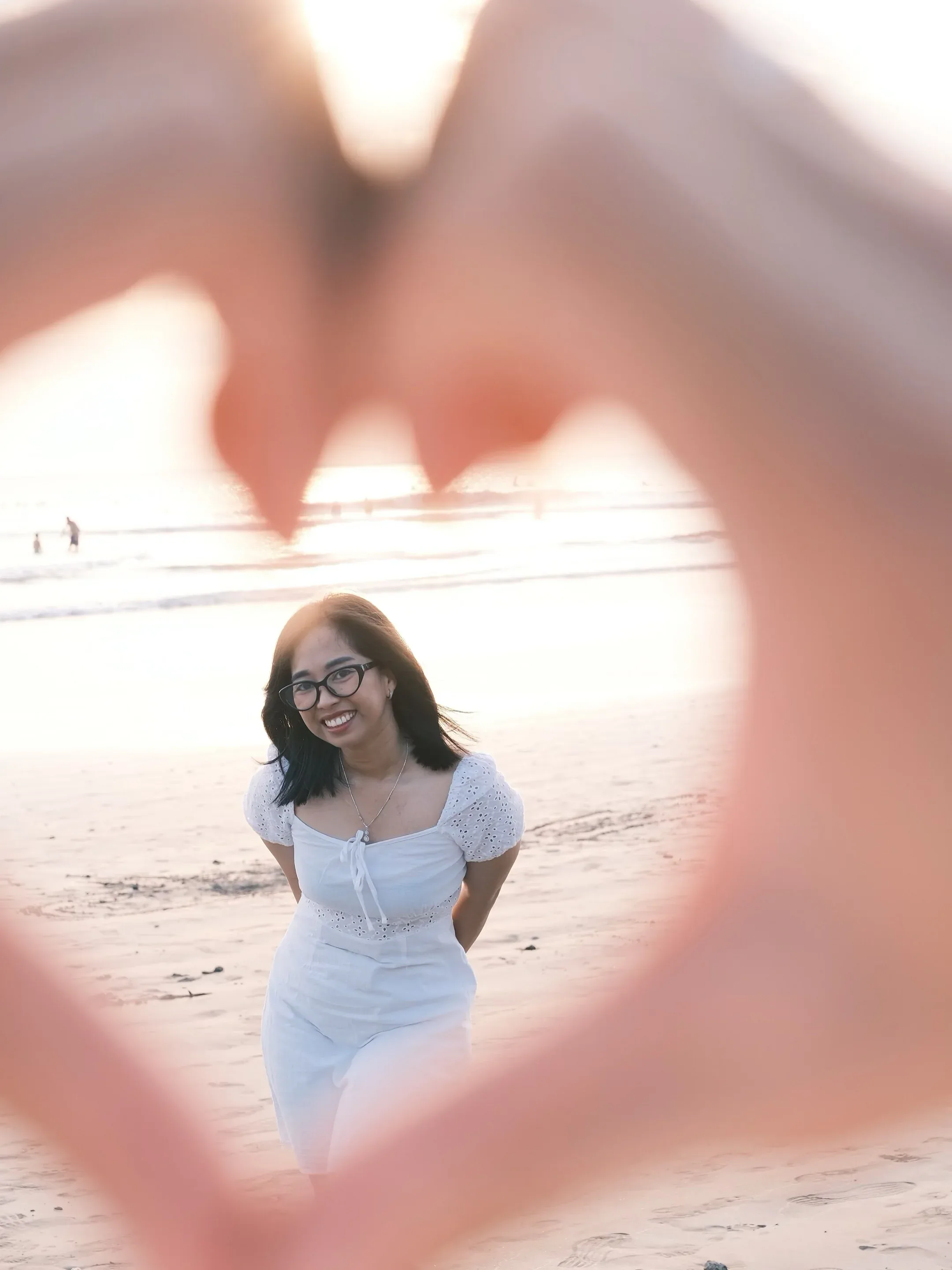 Her standing on the sand, framed by two hands held up in a heart shape in the foreground, sun behind her.