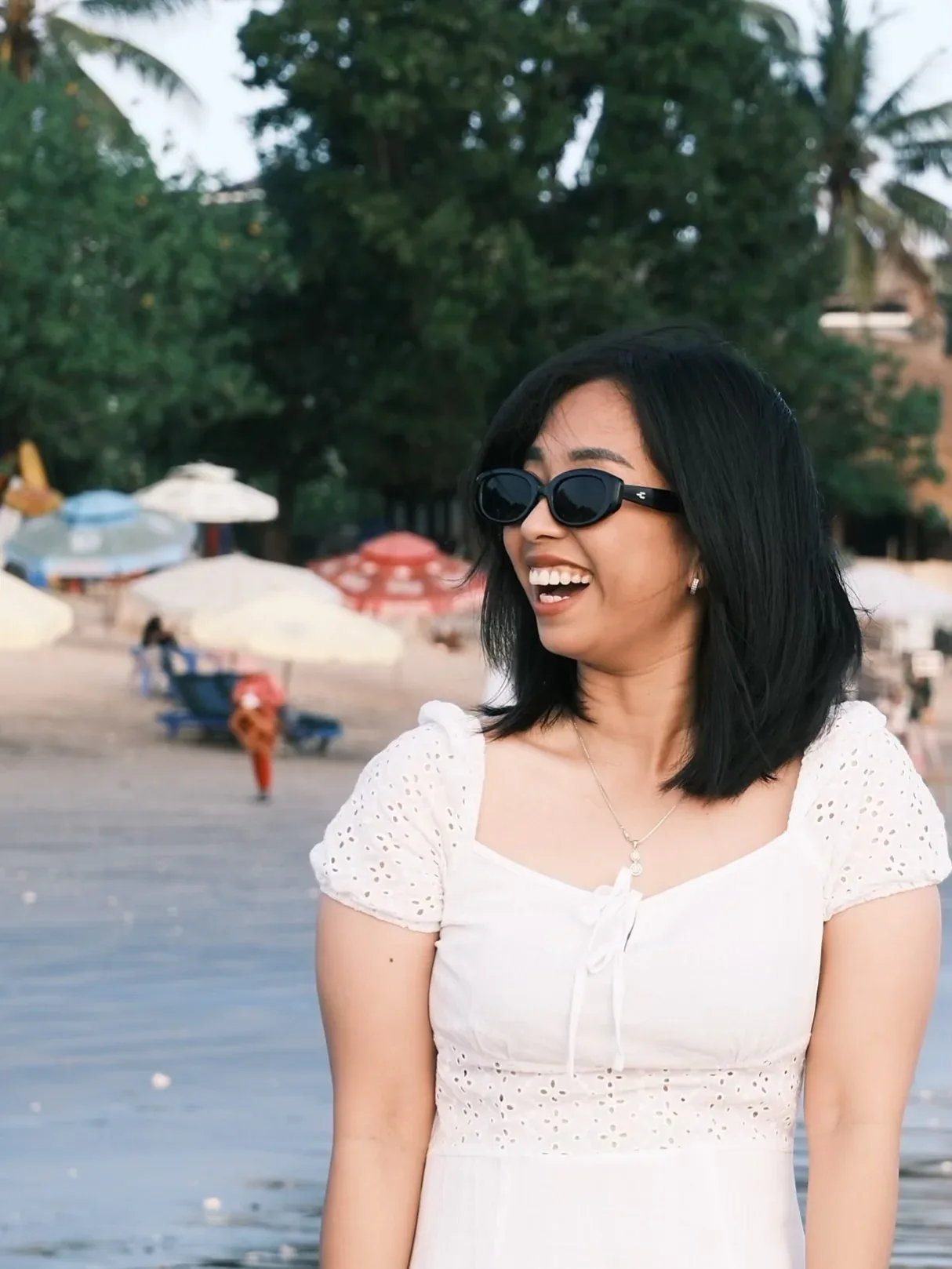 Portrait of Chintya laughing in sunglasses and a white eyelet dress, beach umbrellas and palms behind her.