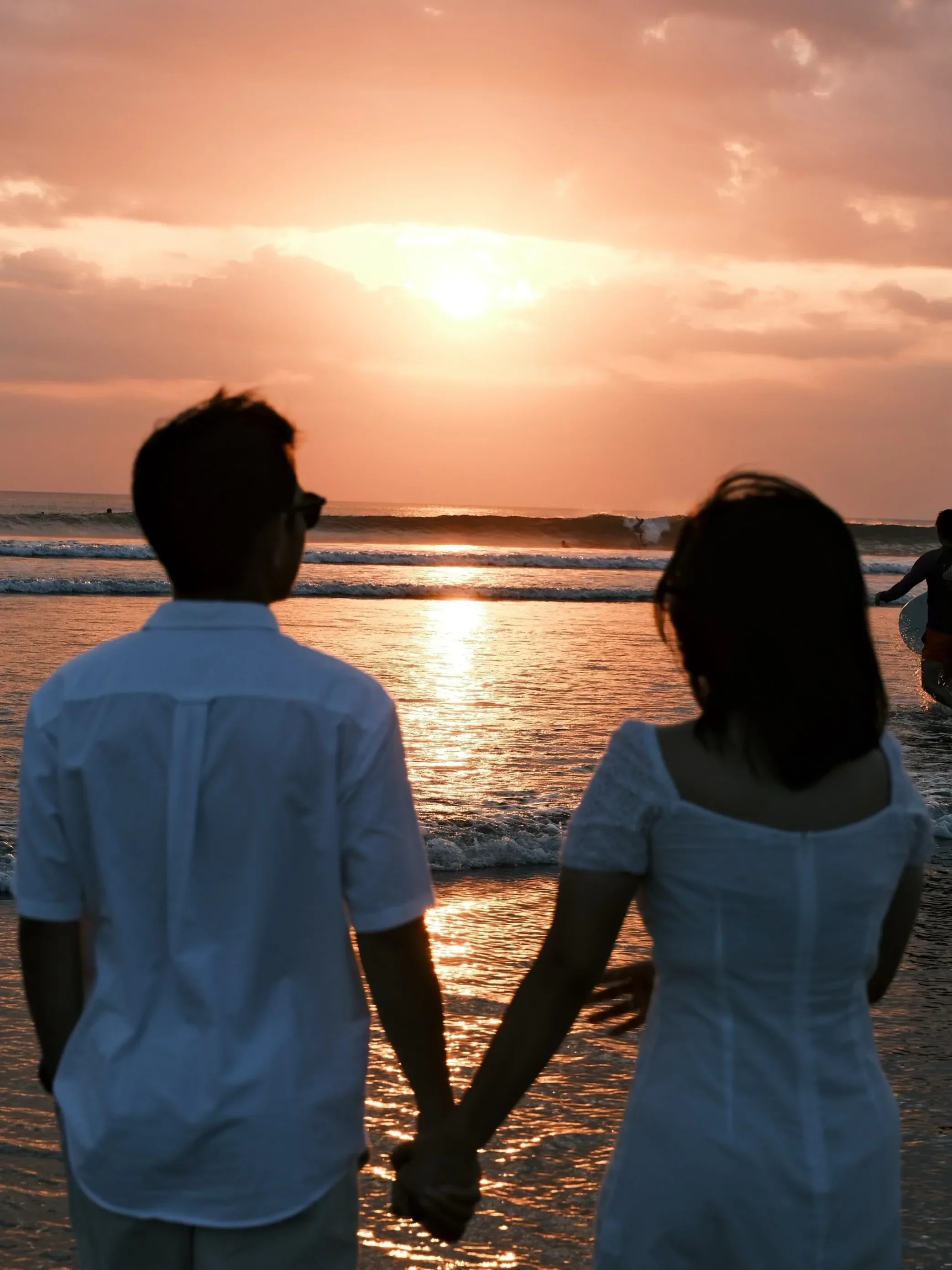 The couple from behind, holding hands, facing an orange sunset over the breaking surf.
