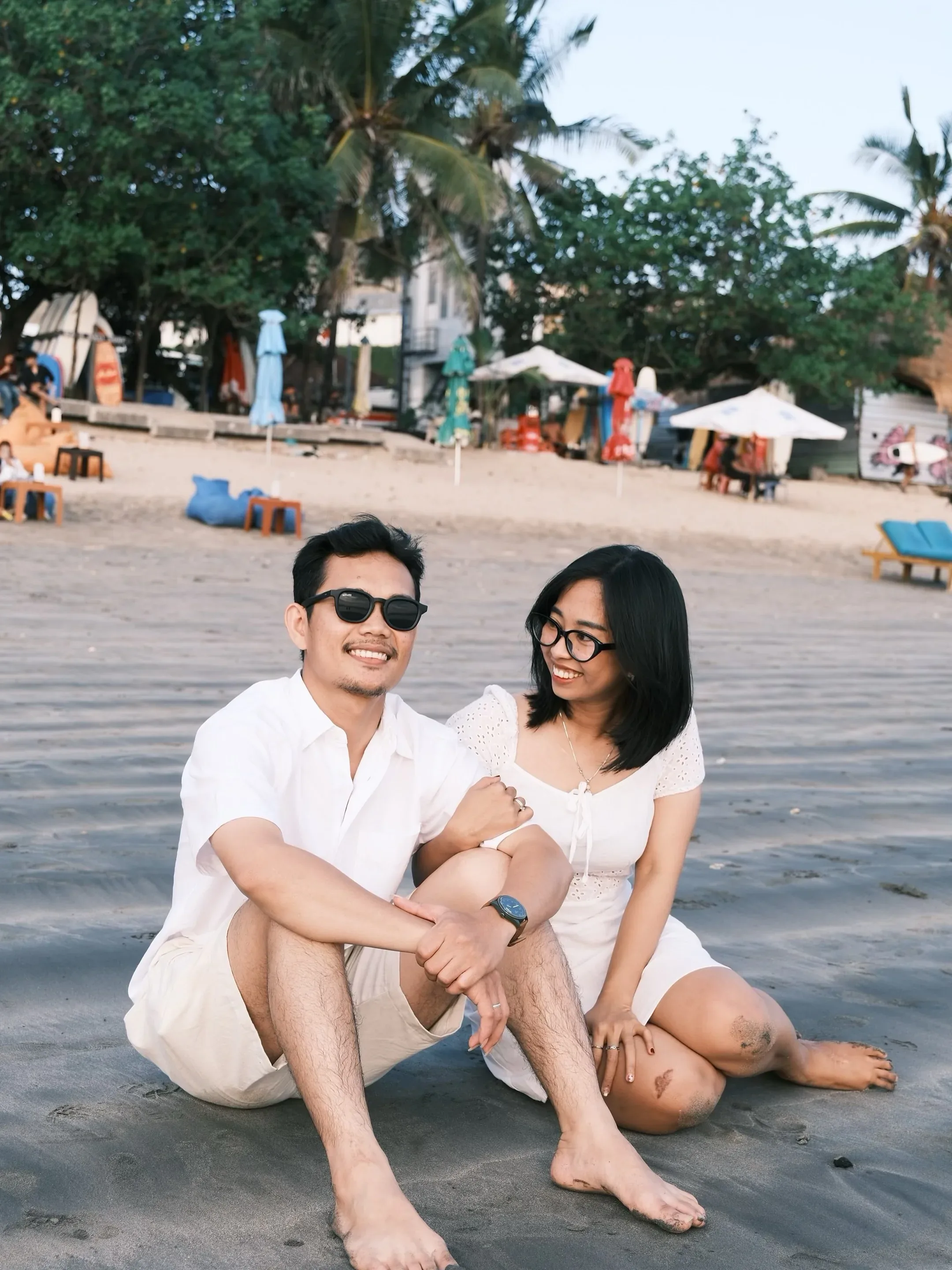 The two of them sitting on the sand in white, smiling at the camera, a beach bar and umbrellas behind.