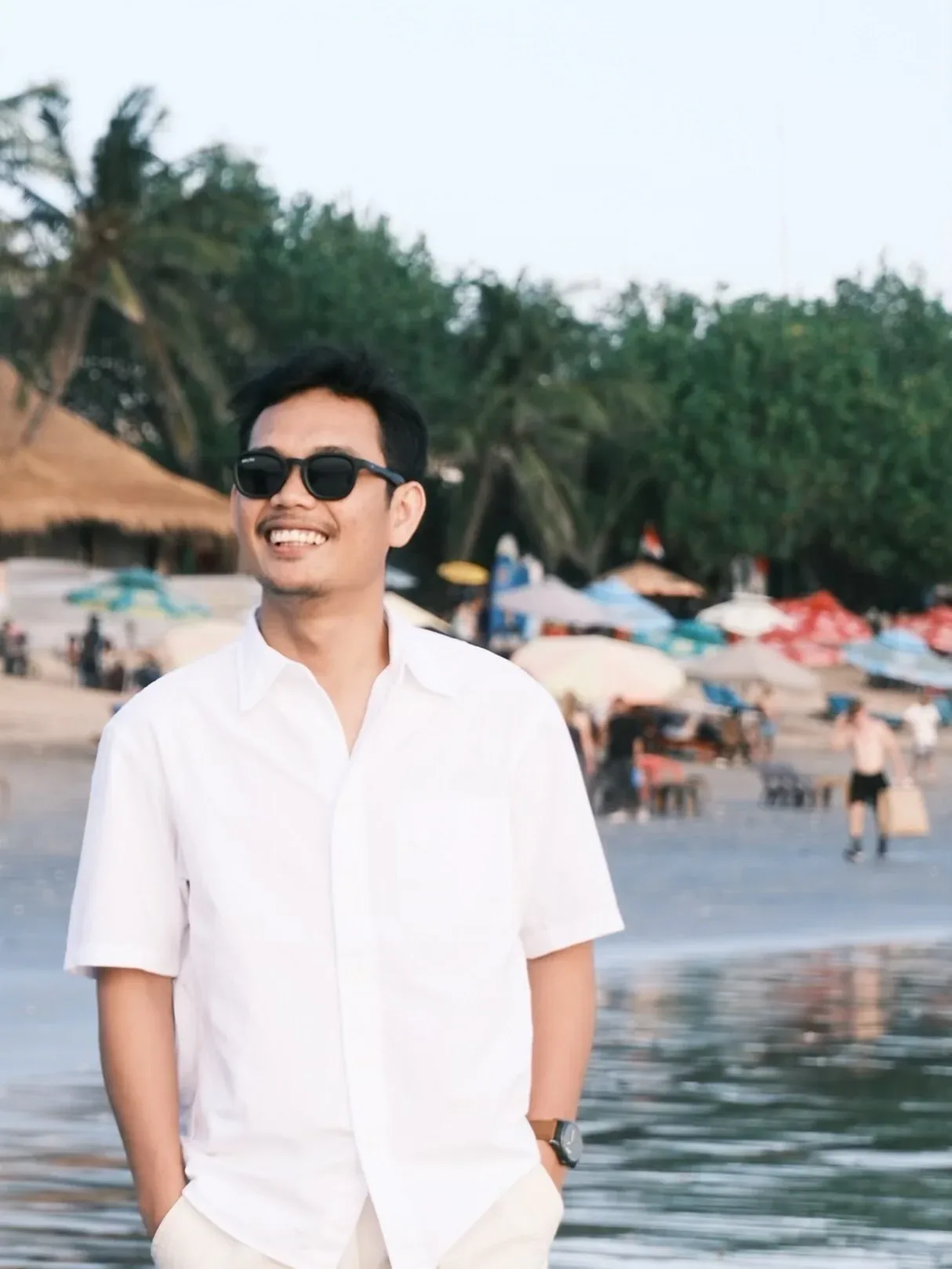 Portrait of Matthew smiling in sunglasses and a white shirt, hands in his pockets, umbrellas and palms behind him.