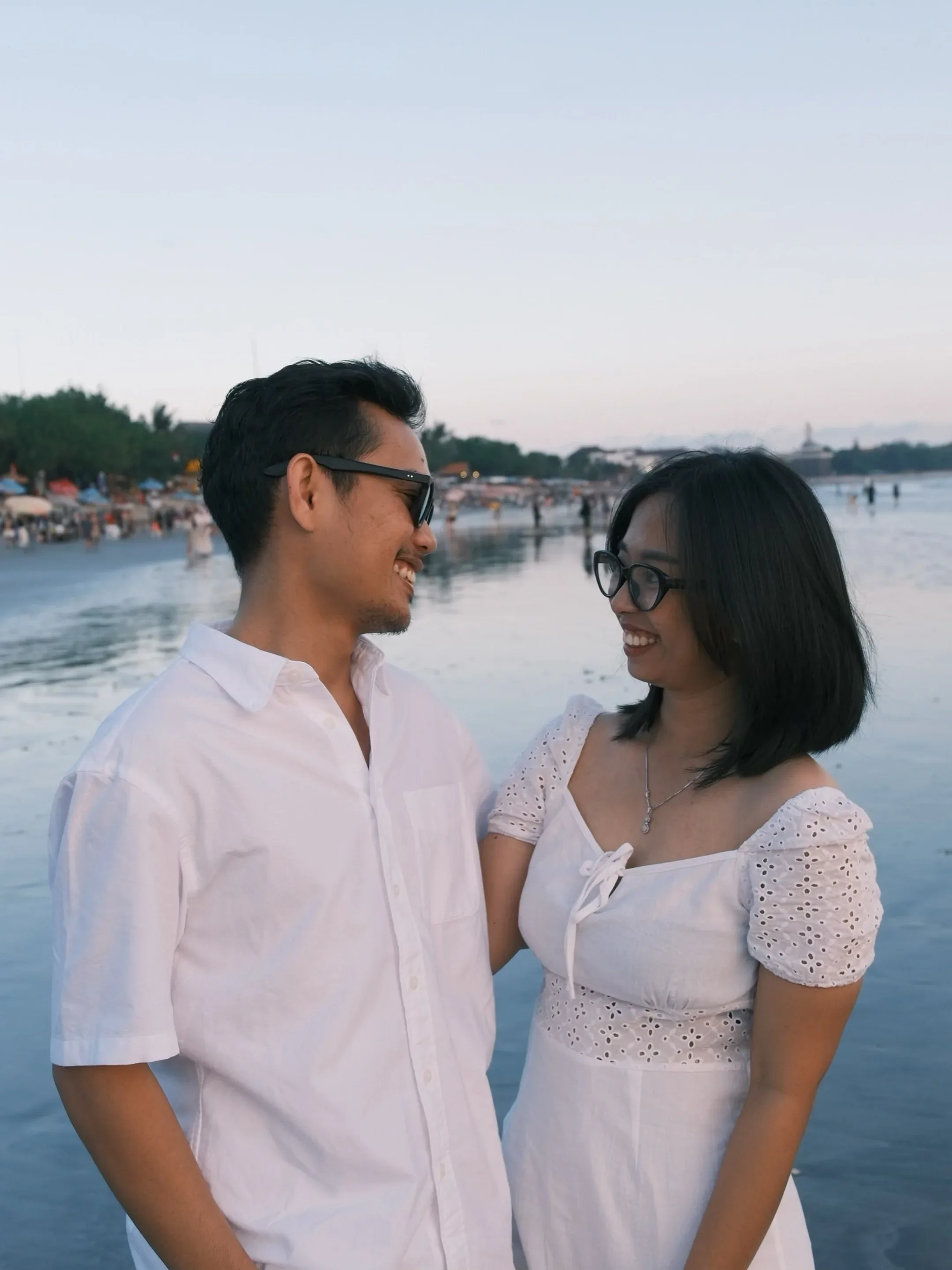 The couple facing each other and laughing, the shoreline and a dusk sky behind them.